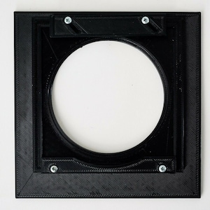 May include: Black square-shaped frame with a round opening in the center. The frame is made of a textured material and has four screws on the edges.