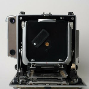 Peut inclure: Un appareil photo moyen format vintage, vu de face avec le dos ouvert. Le corps de l'appareil est argenté et beige, avec des détails noirs et argentés. Le dos ouvert révèle le compartiment à film et les mécanismes internes. L'appareil est conçu pour la photographie professionnelle.