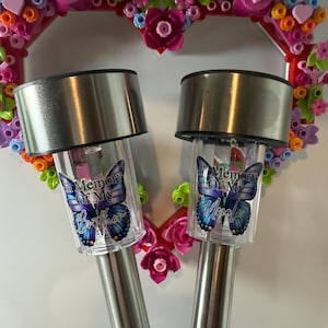 May include: Two solar garden lights with silver tops and butterfly graphics that read "In Memory Of My Brother" and "In Memory Of My Uncle". A heart-shaped wreath with pink and colourful flowers is in the background.