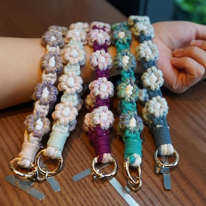 May include: Five handmade macrame bracelets in various colors, including purple, teal, and cream. Each bracelet features a floral design with a gold-colored ring and a silver-colored clasp. The bracelets are displayed on a wrist.