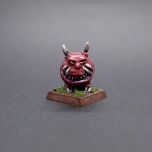 May include: A small, red, painted miniature figurine of a demon with horns, a toothy grin, and yellow eyes. The demon is standing on a brown base with green grass.
