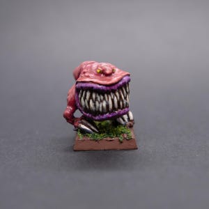 May include: A small, red, painted miniature of a creature with a large, toothy mouth. The creature has yellow eyes and is standing on a brown base with green grass.