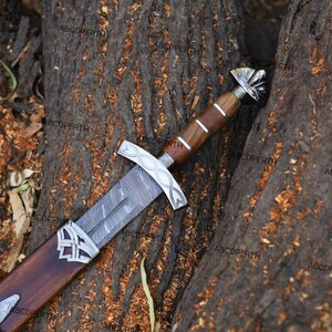 May include: A detailed image of a decorative sword with a wooden handle and sheath. The blade has a patterned design, and the hilt and sheath feature silver-colored accents. The sword rests on a textured, brown surface.