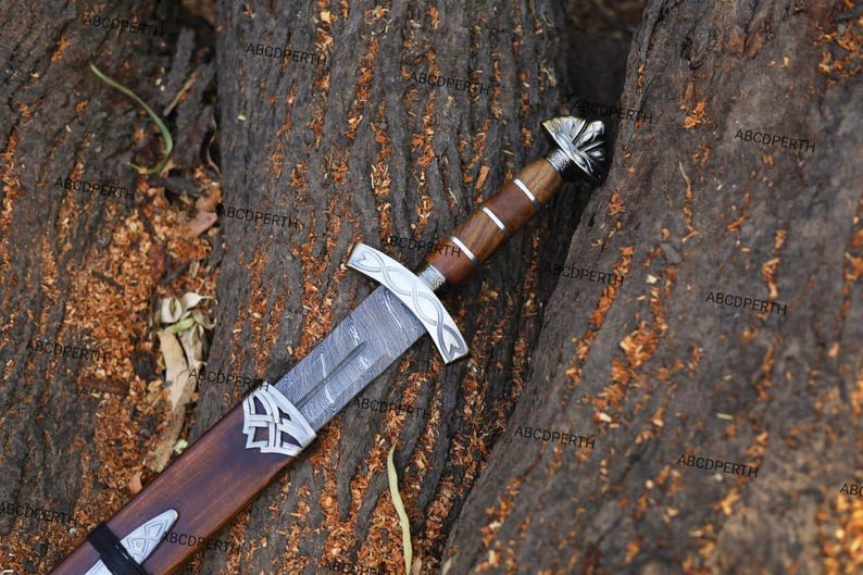 May include: A detailed image of a decorative sword with a wooden handle and sheath. The blade has a patterned design, and the hilt and sheath feature silver-colored accents. The sword rests on a textured, brown surface.