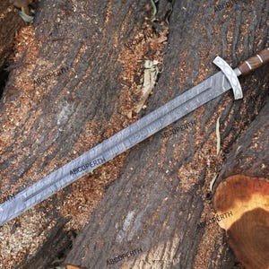 May include: A long, silver sword with a patterned blade and a wooden handle. The sword has a silver crossguard and pommel. The sword is lying on a bed of brown wood.