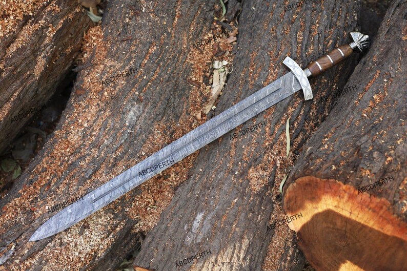 May include: A long, silver sword with a patterned blade and a wooden handle. The sword has a silver crossguard and pommel. The sword is lying on a bed of brown wood.