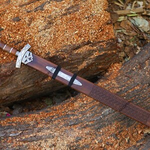 May include: A decorative sword with a wooden handle and sheath, resting on a wooden surface. The sword features a silver crossguard and pommel, with a detailed pattern on the sheath. The blade is not visible.