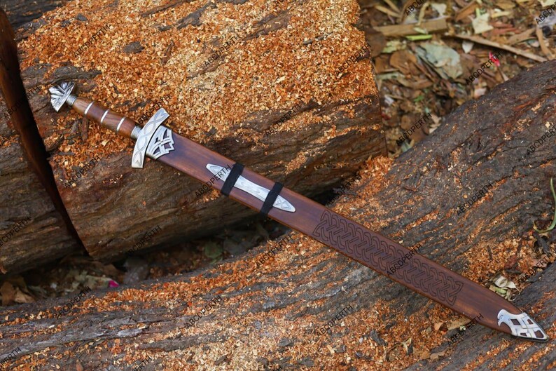 May include: A decorative sword with a wooden handle and sheath, resting on a wooden surface. The sword features a silver crossguard and pommel, with a detailed pattern on the sheath. The blade is not visible.