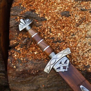 May include: A detailed view of a Viking sword with a wooden handle and sheath. The handle is wrapped with metal bands and features a decorative metal pommel. The blade is housed in a wooden sheath with metal accents. The sword rests on a wooden surface.