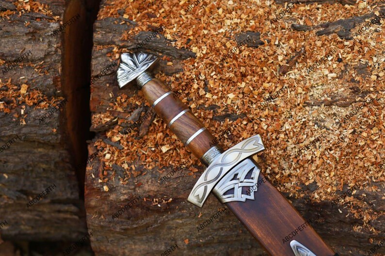 May include: A detailed view of a Viking sword with a wooden handle and sheath. The handle is wrapped with metal bands and features a decorative metal pommel. The blade is housed in a wooden sheath with metal accents. The sword rests on a wooden surface.