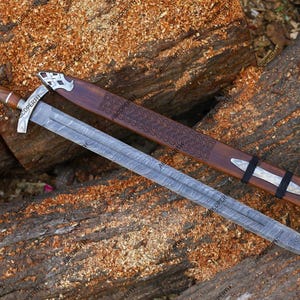 May include: A long, silver sword with a patterned blade and wooden handle rests on a wooden surface. The sword is accompanied by a brown sheath with decorative carvings and silver accents. The handle has a silver crossguard.