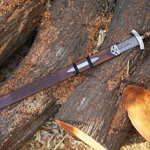 May include: A long, ornate sword with a wooden scabbard rests on a bed of logs. The sword features a silver blade, a detailed hilt, and a brown handle. The scabbard is brown with silver accents and a decorative pattern.