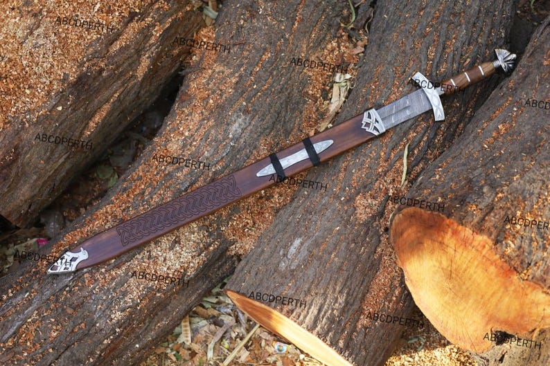 May include: A long, ornate sword with a wooden scabbard rests on a bed of logs. The sword features a silver blade, a detailed hilt, and a brown handle. The scabbard is brown with silver accents and a decorative pattern.