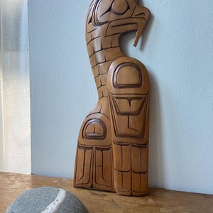 Peut inclure: Sculpture en bois sculpté d'un oiseau au cou incurvé et aux détails complexes. La sculpture est de couleur marron clair et se dresse verticalement. Une pierre grise avec une bande blanche est devant la sculpture.