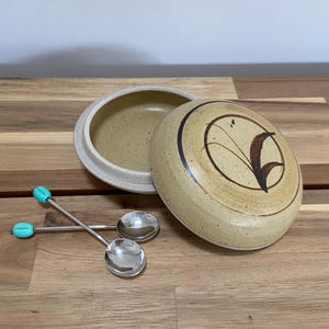 Op de afbeelding: Een ronde, gespikkelde beige keramische schaal met deksel, voorzien van een donkerbruin geschilderd bladmotief op het deksel. Twee kleine zilverkleurige metalen lepels met helder turquoise accenten op de handvatten liggen naast de open schaal.