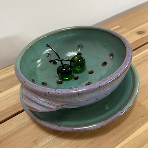 May include: A ceramic berry bowl with a matching saucer, featuring a speckled teal interior and a lavender-toned exterior. The bowl has drainage holes and contains two small, dark green glass cherry ornaments with black stems inside.