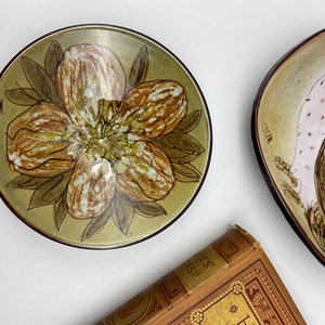 Op de afbeelding: Twee keramische borden met handgeschilderde ontwerpen. Een bord heeft een bruine en groene bloem met witte accenten. Het andere bord heeft een bruine en witte uil met roze accenten.