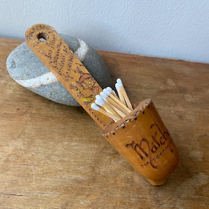 Könnte beinhalten: Ein hellbrauner Holz-Streichholzhalter mit dem Wort "Match" auf der Seite. Er enthält weißspitzige Streichhölzer. Der Griff ist mit Text und einem Cartoon-Bild graviert. Der Halter liegt auf einer Holzoberfläche.