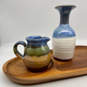 May include: A small, ceramic creamer with a handle and spout, featuring bands of blue, green, and yellow glaze. Beside it, a tall, blue and white ceramic vase sits on a wooden tray. The tray has a natural wood grain finish.