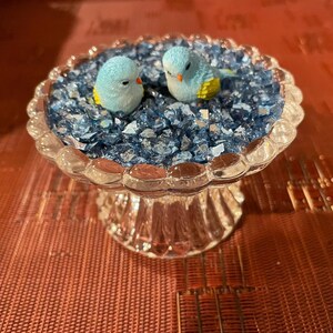 May include: Two small blue bird figurines are perched in a clear glass bird bath filled with blue glass stones.