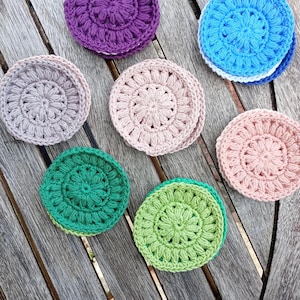 May include: A selection of handmade crochet coasters in shades of purple, blue, green, and pink. The round coasters feature a floral pattern, ideal for protecting surfaces from drink rings. Each coaster is approximately 10 cm in diameter.