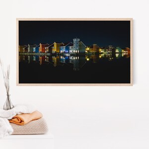 Op de afbeelding: Een nachtzicht van een rij kleurrijke huizen die worden weerspiegeld in een stil waterlichaam. De huizen zijn verlicht met warme gele lichten, wat een gezellige en uitnodigende sfeer creëert.