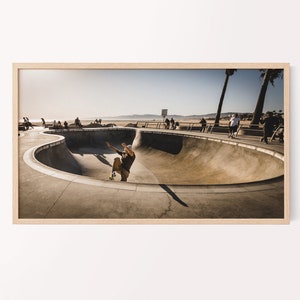 Op de afbeelding: Een skateboarder voert een truc uit in een betonnen skatepark met uitzicht op het strand en palmbomen op de achtergrond.
