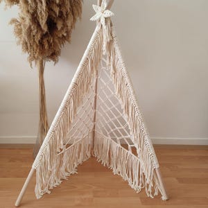 Op de afbeelding: Een handgemaakte tipi-tent in crèmekleur met een macramé-ontwerp. De tent heeft een gefranjerde rand en is versierd met een stervormige versiering aan de bovenkant. De tent wordt ondersteund door houten palen.
