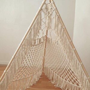 Op de afbeelding: Een crèmekleurige macramé tipi met een houten frame. De tipi heeft een driehoekige vorm, waarbij het macramé de wanden vormt en een decoratief patroon aan de bovenkant. De franje hangt naar beneden vanaf het macramé.
