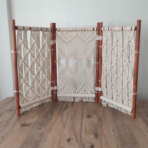 Op de afbeelding: Een macramé roomdivider met drie panelen. Het houten frame is warmbruin en de macramé panelen zijn gebroken wit. Elk paneel heeft een uniek geometrisch patroon. De divider is vrijstaand.