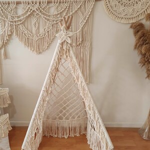 Op de afbeelding: Een crèmekleurige macramé tipi met een gefranjerde rand en een stervormige top. De tipi staat tegen een muur met bijpassende macramé wandkleden en kussens. Een vaas met gedroogd pampasgras voegt een natuurlijke touch toe.