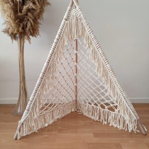 Op de afbeelding: Een crèmekleurige macramé tipi-tent met houten palen. De tent heeft een geweven netontwerp en franjes langs de randen. De tent staat op een houten vloer, met een hoge vaas met gedroogd pampasgras op de achtergrond.
