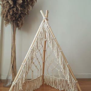 Op de afbeelding: Een crèmekleurige macramé tipi met houten palen. De tipi heeft een geweven netontwerp en franjes aan de onderkant. Op de achtergrond staat een hoge glazen vaas met gedroogd pampasgras. De tipi staat op een houten vloer.