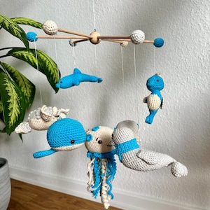 Peut inclure: Un mobile pour bébé avec un hippocampe, un dauphin, une baleine, une méduse et un phoque en crochet bleu et blanc. Le mobile est suspendu à un cadre en bois avec des boules blanches.