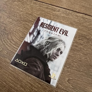 May include: A Resident Evil: Requiem game card featuring a woman with blonde hair. The card has the game title in white text and the PlayStation logo in gold. The background is a dark, blurred image.