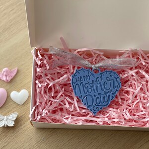 May include: A blue heart-shaped ornament with the text "Happy Mother's Day" inside a white box filled with pink paper shreds. The box is on a wooden surface, with decorative elements including a pink angel, a pink heart, a white heart, and a white butterfly.