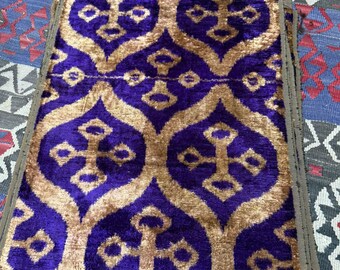 May include: A folded, rectangular textile with a repeating pattern of gold-colored shapes on a deep purple background. The edges are trimmed with a brown border. The textile is resting on a patterned rug.