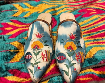 May include: A pair of blue and white patterned mules with floral embroidery in red, yellow, and green. The shoes have pointed toes and brown trim. The background is a colorful patterned fabric in teal, yellow, and pink.