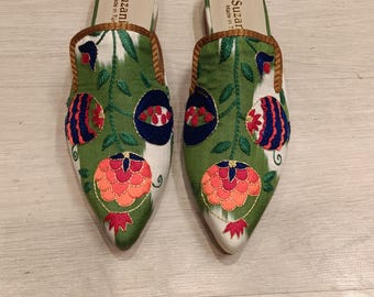 May include: A pair of embroidered green and white slip-on shoes. The shoes feature a pointed toe and are decorated with floral patterns in shades of red, orange, blue, and green. The trim is brown.