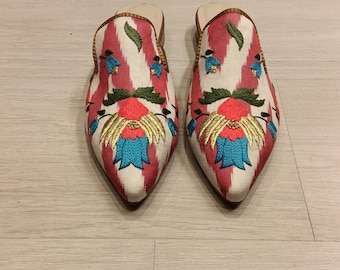May include: A pair of embroidered slip-on shoes. The shoes have a pointed toe and a cream-colored base with a red and white striped pattern. The shoes are decorated with floral embroidery in shades of red, blue, and gold.