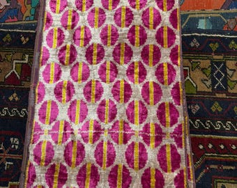 May include: A rectangular textile with a repeating pattern of pink circles with yellow vertical stripes on a silver background. The fabric is folded over, revealing a glimpse of the underside. The textile is placed on a patterned rug.