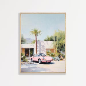 May include: A framed art print depicting a light pink sports car parked in front of a modern white building with a flat roof. A tall palm tree stands beside the structure, surrounded by green foliage and desert plants under a clear blue sky.
