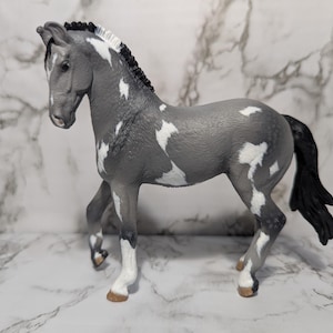 Pode incluir: Estatueta de cavalo de brinquedo cinzento e branco. O cavalo tem uma crina e cauda pretas, com manchas brancas no corpo. O cavalo está em pé, com uma perna levantada. A estatueta é feita de plástico e é uma representação detalhada de um cavalo.