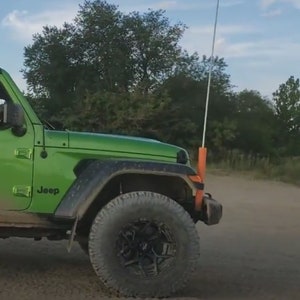 May include: A green Jeep Wrangler with a black bumper and black wheels. The Jeep is parked on a dirt road with trees in the background.