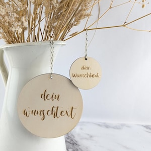 Könnte beinhalten: Zwei runde Holzanhänger mit dem Text "dein Wunschtext" in kursivschrift. Die Anhänger hängen an einer weißen Schnur und befinden sich vor einem weißen Marmor Hintergrund.