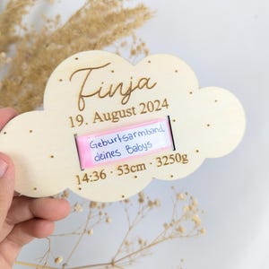 Könnte beinhalten: Ein Holz-Wolken-Andenken mit dem Namen "Finja" und dem Datum "19. August 2024" eingraviert. Ein rosa Band mit dem Text "Geburtsarmband deines Babys" ist an der Wolke befestigt. Das Band enthält auch die Uhrzeit, die Länge und das Gewicht des Babys: "14:36 - 53cm - 3250g".