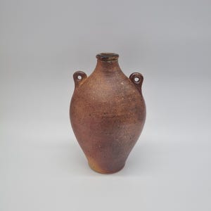 May include: A rustic, brown ceramic vase with two small handles and a narrow neck. The vase has a rounded body and a speckled texture, with color variations from light brown to deep reddish-brown.
