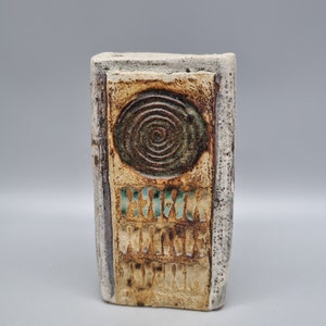 May include: A ceramic vase with a textured surface and a spiral design in brown, green, and white. The vase has the words "HOW WILL YOU SERVE" written on it.