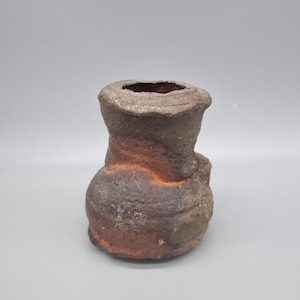 May include: A handmade, rustic ceramic vase with a bulbous base and a cylindrical neck. The vase has a rough, textured surface with a blend of brown, orange, and grey tones. The opening at the top is oval-shaped.