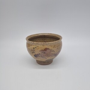 May include: A handmade ceramic bowl with a speckled tan exterior and a dark brown interior. The bowl has a rounded shape with a small, sturdy base. Decorative brown and red accents are visible on the exterior.